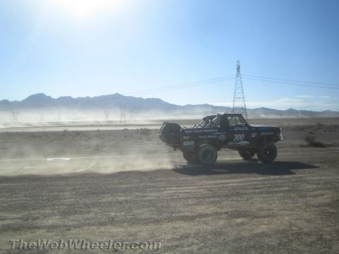 1990 Ford Bronco - Race truck number 300 from West Coast Broncos drives across a dusty desert course beside a large power-line tower.