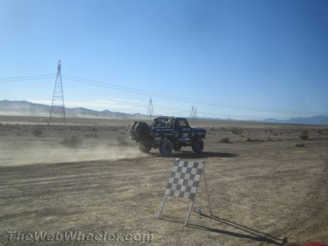 1990 Ford Bronco - off-road race truck drives past a checkered-flag sign on a dusty desert course under a clear sky.