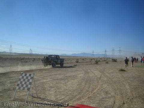 1990 Ford Bronco - dark off-road truck raises dust on a desert course past a checkered-flag sign as people walk nearby.