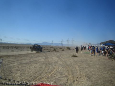 1990 Ford Bronco - dark off-road truck drives toward a crowd gathered under a canopy at a dusty desert event with power lines behind.