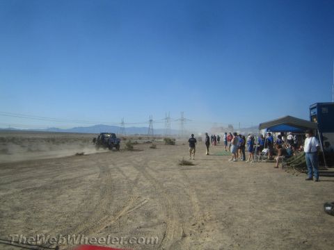 1990 Ford Bronco - distant off-road vehicle raises dust near a crowd and canopy tents at a desert race event under a blue sky.