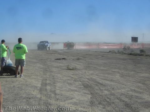 1990 Ford Bronco - off-road truck drives through heavy dust along an orange-fenced desert course as green-shirted spectators watch.