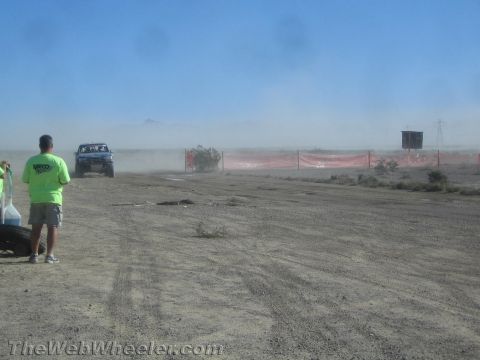 1990 Ford Bronco - blue off-road truck drives along an orange-fenced desert course amid heavy dust as a green-shirted spectator watches.