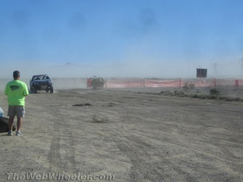1990 Ford Bronco - blue off-road truck kicks up dust along an orange-fenced desert course as a spectator in green looks on.