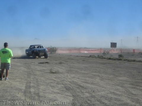 1990 Ford Bronco - blue prerunner race truck rounds a dusty desert course past an orange snow-fence barrier as a spectator looks on.
