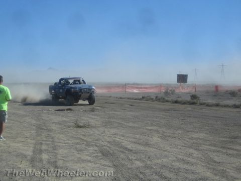 1990 Ford Bronco - blue off-road truck corners on a hardpack desert track lined with orange fencing, trailing a cloud of dust.