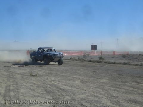 1990 Ford Bronco - blue prerunner truck powers through a dusty desert course with power-line towers visible in the haze behind.