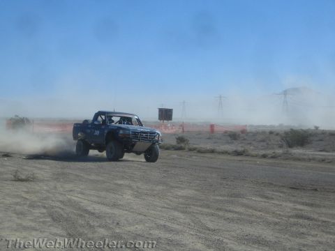 1990 Ford Bronco - blue off-road race truck slides across a dusty desert track, throwing up a wide dust trail near orange course fencing.