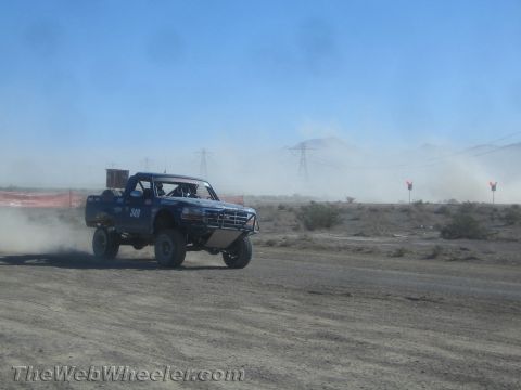 1990 Ford Bronco - Blue race truck number 349 charges along a desert course, distant mountains and power lines visible through the dust.