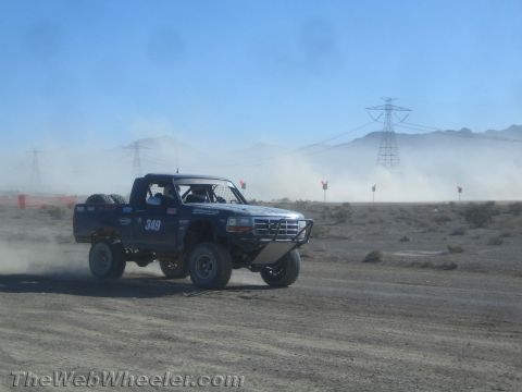 1990 Ford Bronco - Blue prerunner truck number 349, carrying spare tires in its bed, races across a desert track past a transmission tower.