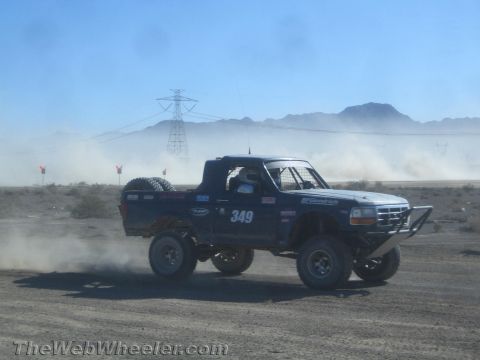 1990 Ford Bronco - dark blue prerunner truck numbered 349 with a white cab roof speeds across a desert course past a power-line tower.