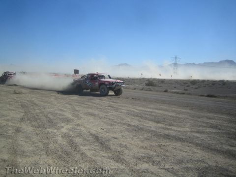 1990 Ford Bronco - red and white off-road race truck kicks up a large dust cloud as it speeds along a desert track with another vehicle behind.