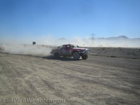 1990 Ford Bronco - red and white prerunner truck corners hard on a desert course, throwing up a thick wall of dust.