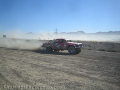 1990 Ford Bronco - red and white off-road race truck with a checkered-flag livery slides across a desert track in a cloud of dust.
