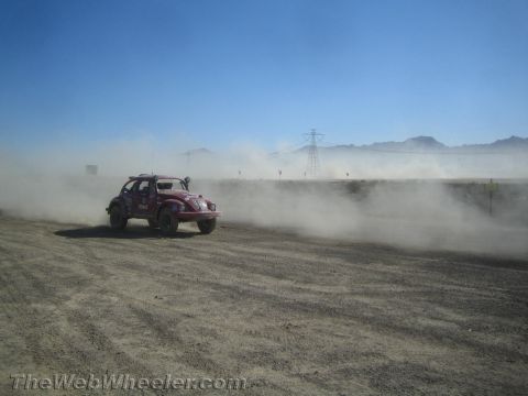 1990 Ford Bronco - dark red Volkswagen Beetle-based Baja Bug races through heavy dust on a desert track with mountains behind.