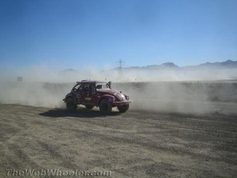1990 Ford Bronco - red Baja Bug with Tecate sponsorship corners on a dusty desert course, mountains and power lines in the distance.