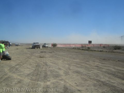 1990 Ford Bronco - Two white vehicles race along a dusty desert track past orange fencing while spectators watch from the left.