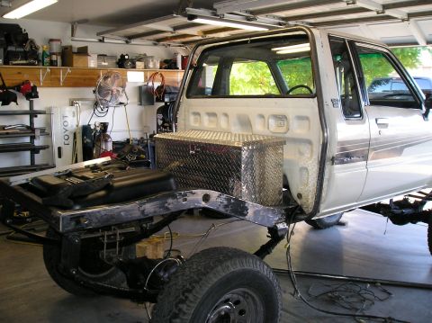 1986 Toyota Hilux - Silver Toyota pickup cab sitting beside its stripped frame in a home garage, mid body-off rebuild with diamond-plate box.