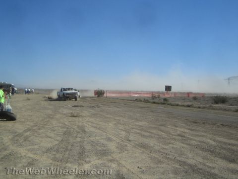 1990 Ford Bronco - white off-road truck speeds along a dusty desert course past orange fencing as spectators watch from the left.