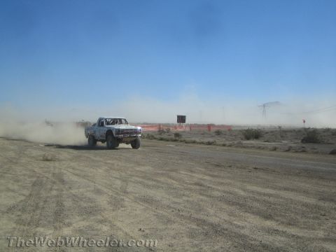 1990 Ford Bronco - white off-road truck speeds along a dusty desert track lined with orange fencing, power-line towers in the haze.