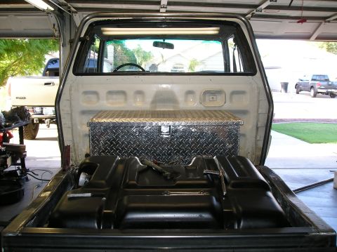 1986 Toyota Hilux - Looking forward into the empty bed of a white pickup with a black steel fuel tank laid flat on the bed floor and hand tools resting on it,…