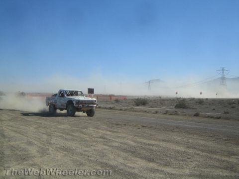 1990 Ford Bronco - white prerunner truck races across a dusty desert course with distant mountains and power-line towers behind.