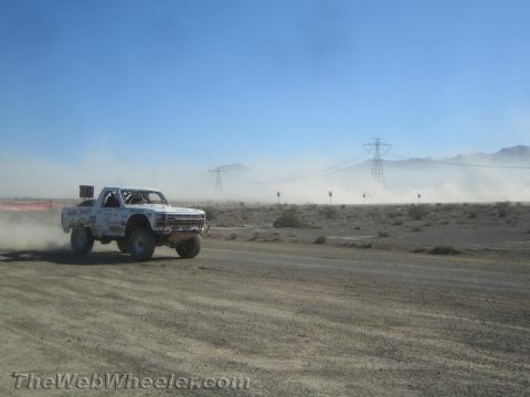 1990 Ford Bronco - White off-road race truck number 302 speeds across a dusty desert course with distant power-line towers and mountains.
