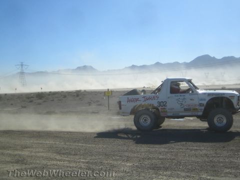 1990 Ford Bronco - white off-road race truck marked 302 races along a dusty desert track beneath power lines and distant mountains.