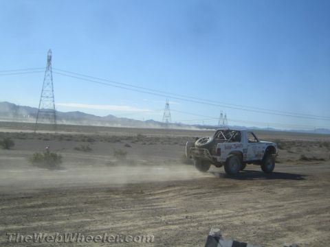 1990 Ford Bronco - white off-road race truck trails dust as it speeds down a desert course past high-voltage transmission towers.