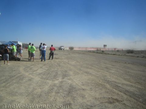 1990 Ford Bronco - Spectators stand at the edge of a desert race course watching a vehicle raise dust in the hazy distance.