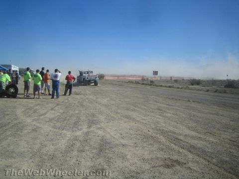 1990 Ford Bronco - group of spectators watches a light-colored off-road race vehicle kick up dust as it approaches on a desert track.