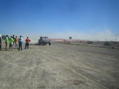 1990 Ford Bronco - off-road race truck trails a plume of dust along a desert course as a line of spectators looks on.