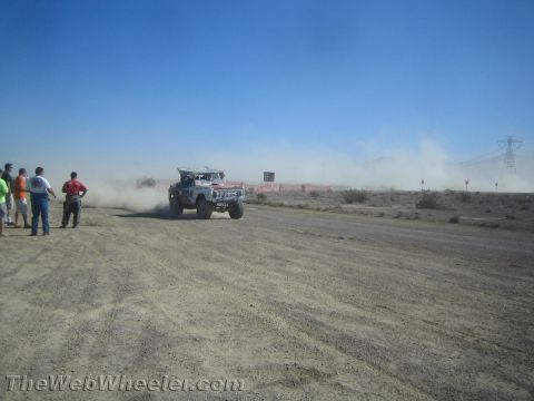 1990 Ford Bronco - white off-road race vehicle raises dust on a desert track while spectators watch from the roadside.