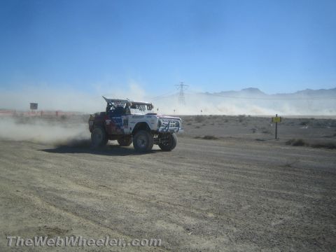 1990 Ford Bronco - white tube-frame off-road race vehicle throws up dust as it rounds a desert course near transmission towers.