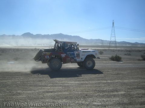 1990 Ford Bronco - Off-road race truck number 305 speeds across an open desert course with mountains and a transmission tower behind.