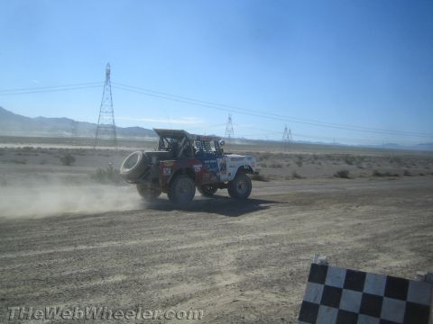 1990 Ford Bronco - white off-road race vehicle carrying a spare tire kicks up dust on a desert course near a checkered marker.