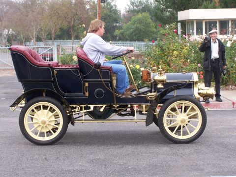 1990 Ford Bronco - Man drives an early 20th-century automobile with a passenger standing beside him.