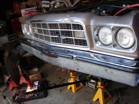 1973 Ford Ranchero - Front end of a silver-tan 1970s Ford up on yellow jack stands inside a cluttered garage, showing quad round headlights in chrome bezels, a…