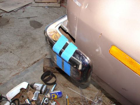 1973 Ford Ranchero - Corner of a chrome bumper on a tan vehicle in a garage, wrapped with strips of blue painter's tape and propped on a wood block during repai…