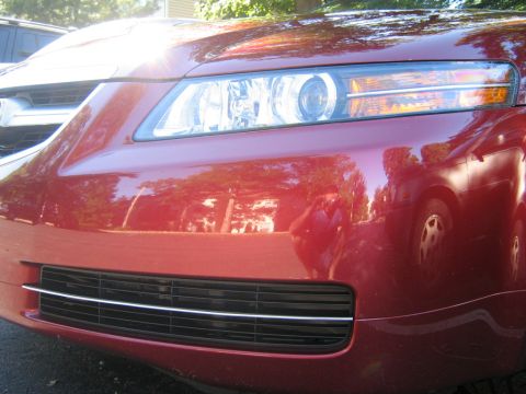 1990 Ford Bronco - Close-up of a red car's front end, showing the grille and headlight, with reflections of trees and another vehicle.
