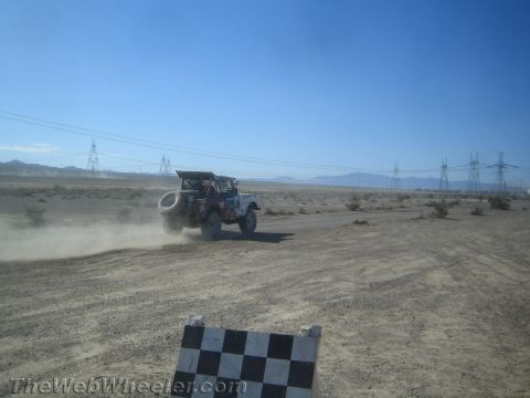 1990 Ford Bronco - off-road race vehicle speeds across a wide desert course past a checkered marker and rows of transmission towers.