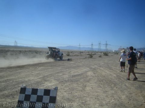1990 Ford Bronco - off-road race vehicle trails dust across the desert while spectators gather near a checkered course marker.