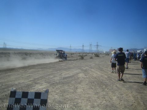 1990 Ford Bronco - Spectators walk along a desert race course as an off-road vehicle raises a trail of dust in the distance.