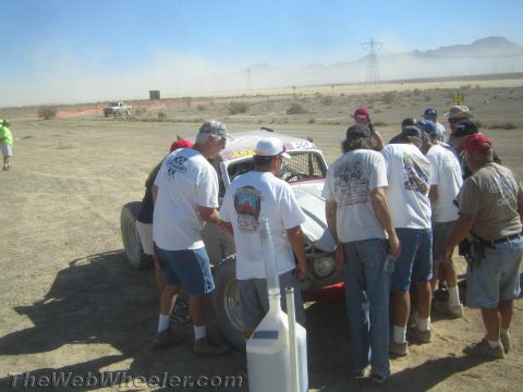 1990 Ford Bronco - crew in matching white shirts gathers around a race car in a dusty desert pit area.