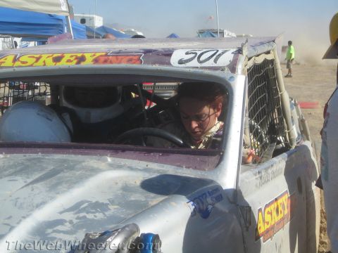 1990 Ford Bronco - person sits behind the wheel in the roll-caged cockpit of a battered silver race car marked 501 in a desert pit.