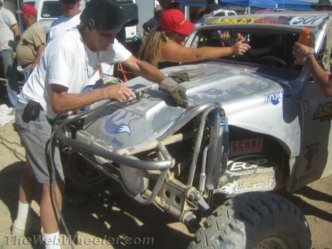 1990 Ford Bronco - Crew members work on the front suspension and windshield of a silver race car during a desert pit stop.