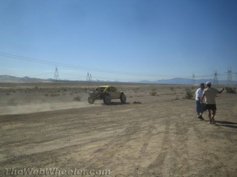 1990 Ford Bronco - yellow off-road race buggy speeds across the desert in the distance while two spectators watch from the trackside.
