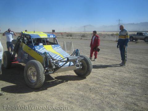 1990 Ford Bronco - yellow and blue tube-frame off-road buggy numbered 1212 sits parked on a desert course as people stand nearby.
