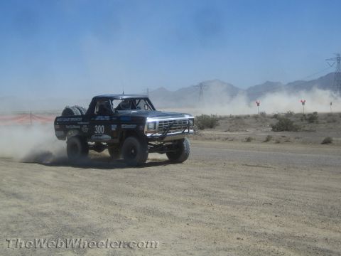 1990 Ford Bronco - black off-road race truck marked 300 powers through a dusty desert course past red course markers.