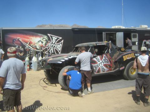 1990 Ford Bronco - crew works on a black graphic-wrapped off-road race truck parked beside a team trailer in the desert pits.